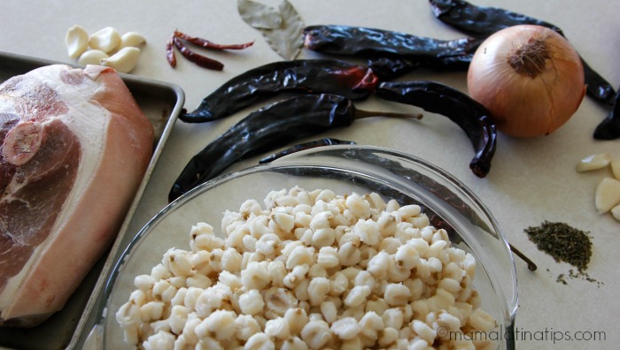 Chiles guajillos, ajos, chiles de árbol, un puñito de orégano, maíz pozolero y carne de puerco en una charola sobre una superficie blanca. 