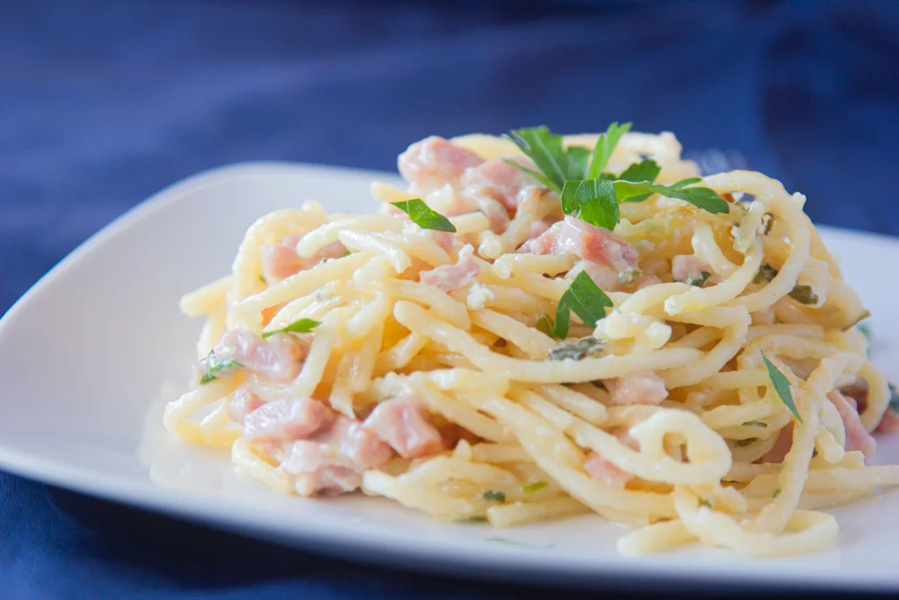 espagueti con jamón, perejil y crema sobre un plato blanco y mantel azul