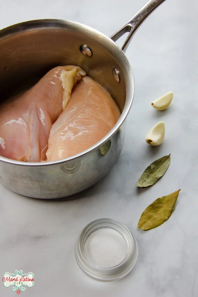Pollo crudo en una olla sobre una mesa blanca, junto a dos ajos, dos hojas de laurel y sal