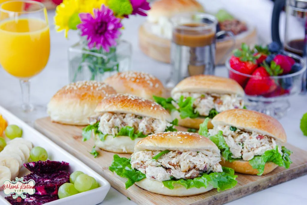 Sándwiches de ensalada de pollo sobre una mesa de brunch. Con jugo de naranja, fruta fresca, café y flores.