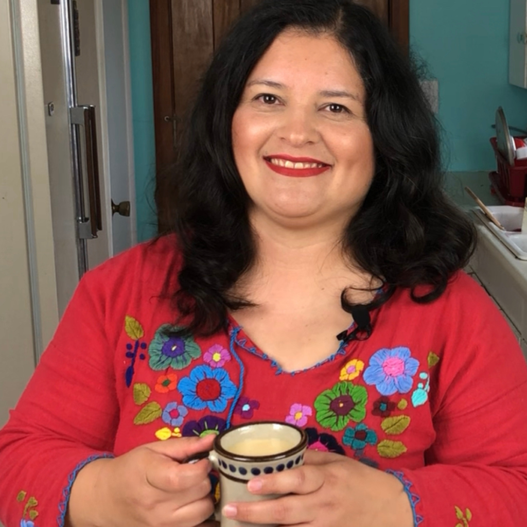 Silvia Martinez from mamalatinatips with long dark hair, wearing a red floral embroidered top, smiles while holding a polka dot mug in a kitchen.