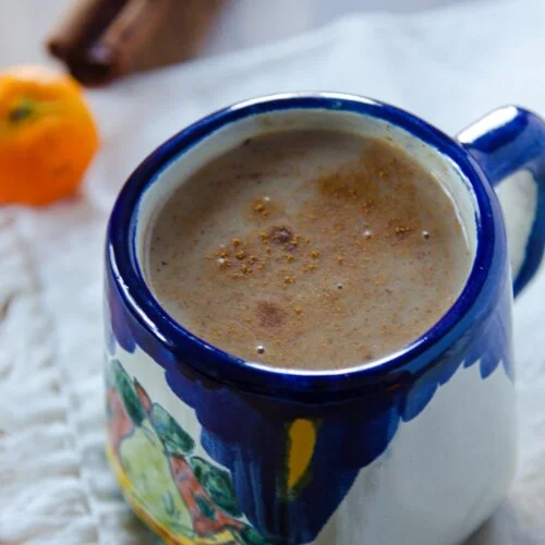 Mexican ceramic cup with atole on top of a fabric napkin. A cinnamon stick next to it.