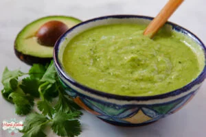 A Mexican ceramic bowl with avocado salsa. There is a bunch of cilantro and a half avocado on the left side of it.
