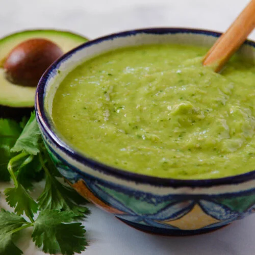 A Mexican ceramic bowl with avocado salsa. There is a bunch of cilantro and a half avocado on the left side of it.