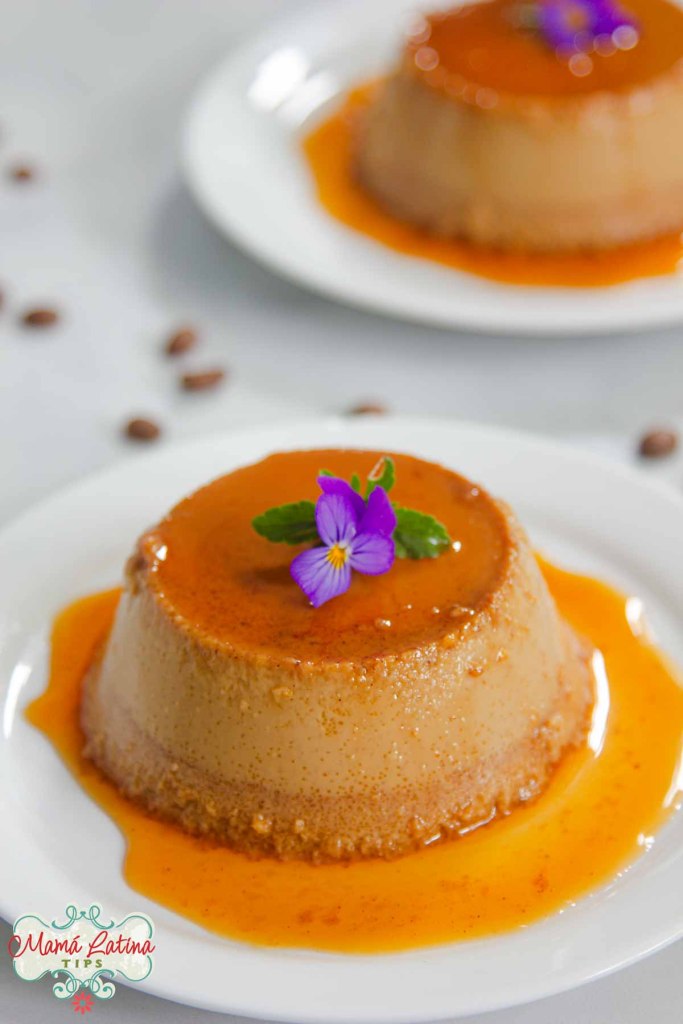 A coffee flan garnished with a purple flower, served on a white plate with a caramel drizzle.