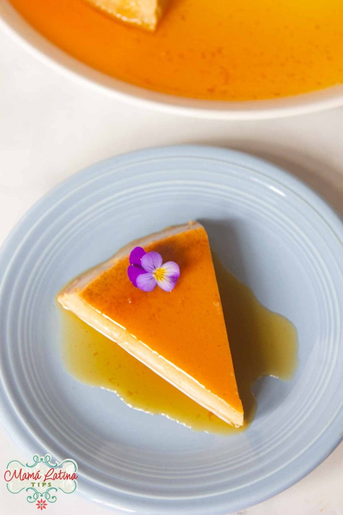 A slice of coffee flan topped with a small purple flower, served on a blue plate with caramel sauce.