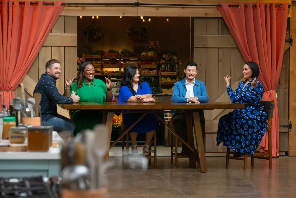Chef Tim Hollingsworth, Chef Tiffany Derry, Silvia from Mama Latina Tips, Chef Francis Lam and Chef Alexandra Ramos sit at a wooden table in a studio kitchen filming The Great American Recipe show, engaged in conversation, with shelves of fresh produce visible in the background.