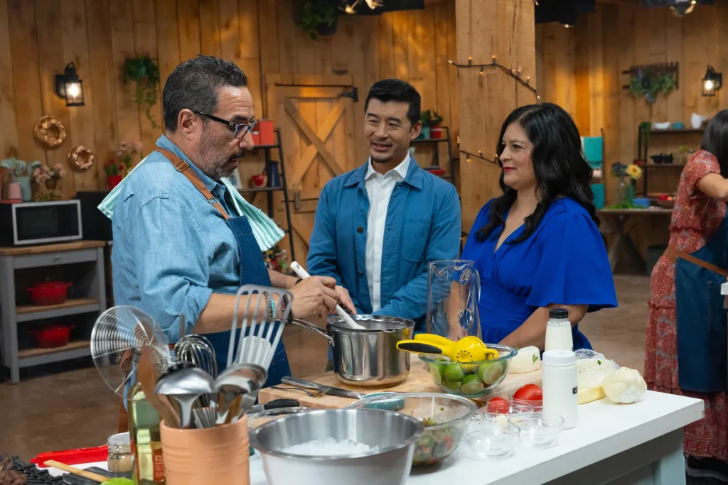 Chef Francis Lam and Silvia Martinez (winner of The Great American Recipe show) stand in a kitchen studio, talking to Jon one of the homecooks participating on the third season of the show while Jon stirring a pot on his cooking station. 